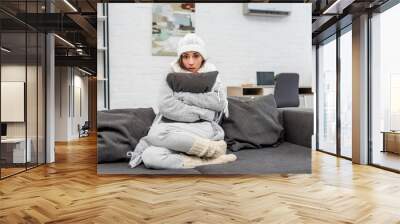 freezed young woman in warm clothes sitting on couch and hugging cushion at home Wall mural