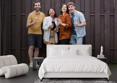 full length of happy interracial friends holding glasses with wine while standing near fence on backyard Wall mural