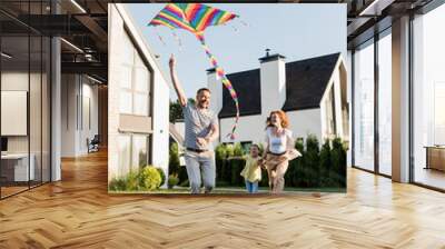 Full length of happy mother and daughter running near father flying kite on lawn near houses Wall mural