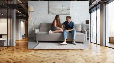 full length of young and happy couple sitting on couch and looking at each other in living room. Wall mural