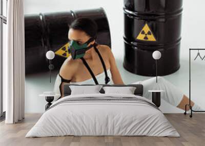 futuristic african american woman in safety mask with gun near radioactive waste barrels on white background Wall mural