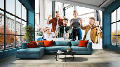 group of young successful entrepreneurs celebrating victory and showing thumbs up at office and looking at camera Wall mural