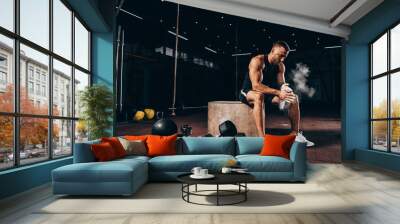 handsome athletic man sitting on cube with gym equipment around and clapping hands with talc before workout Wall mural