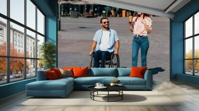 handsome boyfriend in wheelchair and girlfriend with coffee in paper cup having walk on street Wall mural