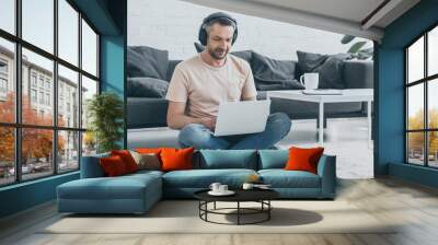 handsome man in headphones sitting on floor with crossed legs and using laptop Wall mural