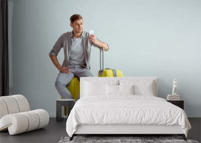 handsome man sitting on yellow suitcase and using smartphone on grey background, travel concept Wall mural