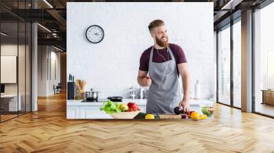 handsome smiling bearded man in apron listening music in earphones and looking away while cooking in kitchen Wall mural