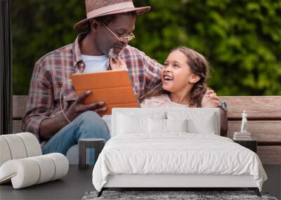 happy african american grandchild and her grandfather listening music on digital tablet while sitting on bench in park Wall mural