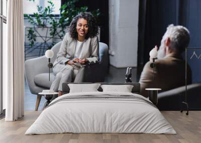 happy african american journalist gesturing while talking with blurred businessman sitting in armchair during interview Wall mural