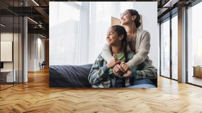 happy african american mother and daughter sitting on couch and looking through window at home Wall mural