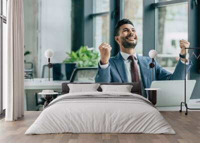 happy businessman looking up and showing winner gesture while sitting at workplace Wall mural