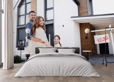 Happy family with daughter hugging and looking at camera near sign with sold lettering and house Wall mural