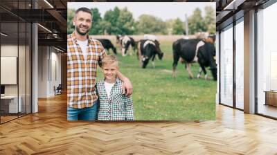 happy father and son smiling at camera while standing near grazing cattle at farm Wall mural