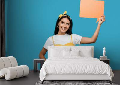 happy girl holding orange speech bubble and standing with hand on hip isolated on blue Wall mural