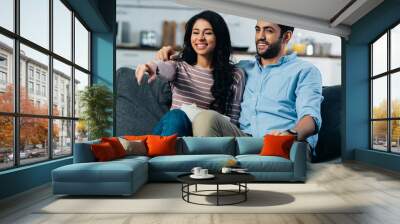 happy latin woman pointing with finger near husband while watching tv at home with bowl of popcorn Wall mural