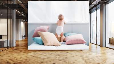 happy little baby in diaper standing on bed with lot of pillows and looking through window Wall mural