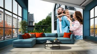 happy man piggybacking daughter near cheerful wife and house Wall mural