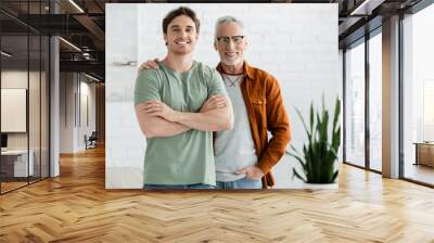 happy mature father in eyeglasses hugging cheerful young son posing with crossed arms at home Wall mural