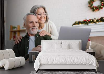 happy middle aged woman in glasses hugging husband and looking at laptop during video call on christmas day Wall mural