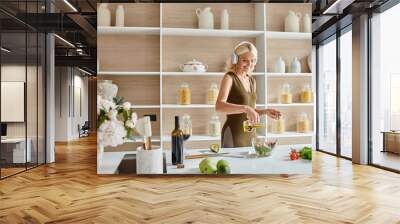 happy middle aged woman in wireless headphones making salad near bottle and glass of red wine Wall mural