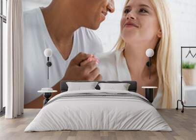 happy multiethnic couple in white sleepwear holding hands and looking at each other in bedroom Wall mural