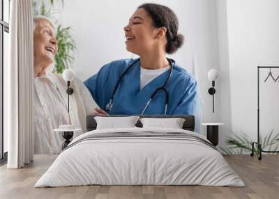 happy multiracial nurse in uniform laughing with retired woman at home, banner. Wall mural