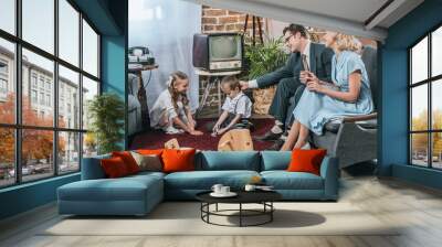 happy parents sitting on sofa and looking at little kids playing with domino tiles at home, 1950s style Wall mural