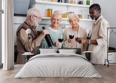 happy retired man holding bottle with wine near joyful multicultural friends in kitchen Wall mural