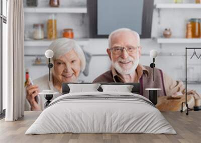 happy senior man adding sliced vegetables into bowl with salad near retired wife with smartphone Wall mural