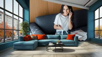 happy woman talking on smartphone and working with business documents on sofa during self isolation Wall mural