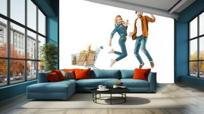 happy young couple jumping near shopping trolley full of products in paper bags isolated on white Wall mural