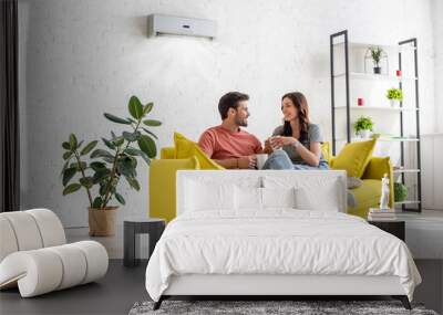 happy young couple talking and holding cups while sitting on yellow sofa under air conditioner at home Wall mural