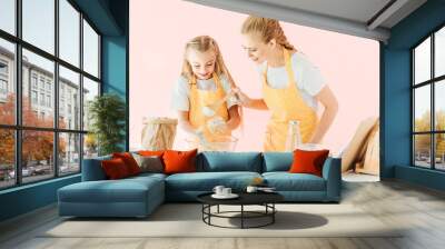 happy young mother and adorable little daughter in yellow aprons making dough together isolated on pink Wall mural