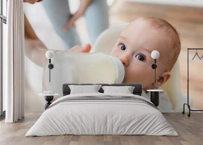 high angle view of toddler boy drinking milk from baby bottle near blurred mom. Wall mural