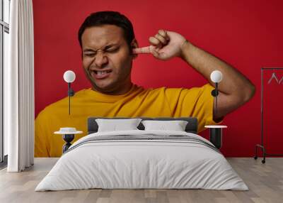 indian man in yellow t-shirt closing eyes and picking his ear with finger on red backdrop, grimace Wall mural