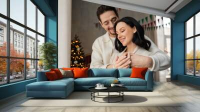 joyful man hugging wife in cozy home wear near blurred decorated Christmas tree, winter holidays Wall mural