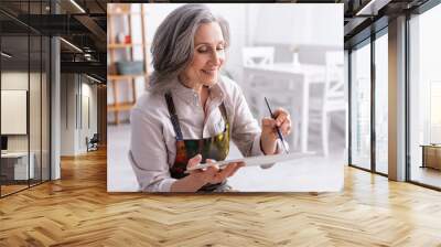 joyful middle aged woman holding paintbrush and palette with colorful paints Wall mural