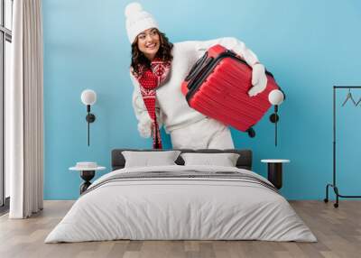 joyful young woman in winter outfit carrying red suitcase on blue Wall mural