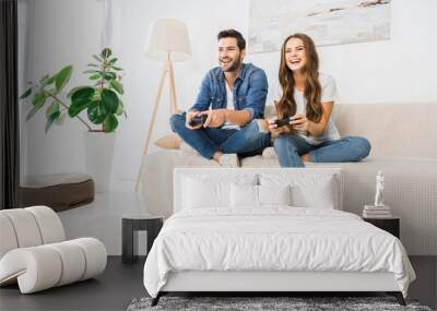 laughing couple playing video game by joysticks on sofa at home Wall mural