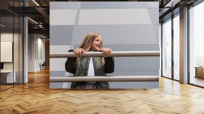 low angle view of cheerful preteen girl in bomber jacket leaning on metallic handrails near mall. Wall mural