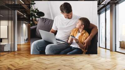 man with laptop looking at adorable son holding credit card Wall mural