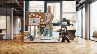 manager in casual attire pointing at analytics on flip chart recording video blog on digital camera Wall mural