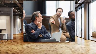 Mature businessman with laptop sitting near multiethnic colleagues in conference room during seminar Wall mural