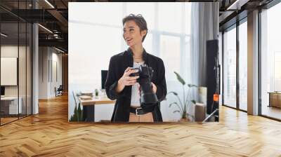 merry female photographer in casual clothes with camera in her hands smiling and looking away Wall mural