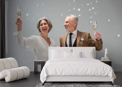 middle aged groom and happy bride in white dress holding glasses of champagne near falling confetti on grey Wall mural