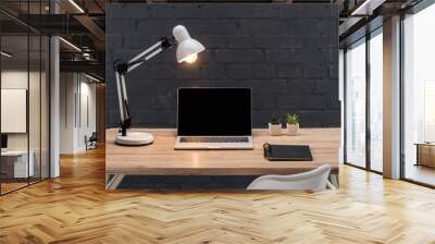 modern workplace with laptop with blank screen, plants, bright lamp and notebook on wooden table near black brick wall Wall mural