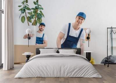 mover holding plant and looking at colleague wrapping cardboard box with scotch tape in apartment Wall mural