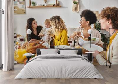 multicultural friends having delicious dinner while gathering on Thanksgiving, women calming child Wall mural