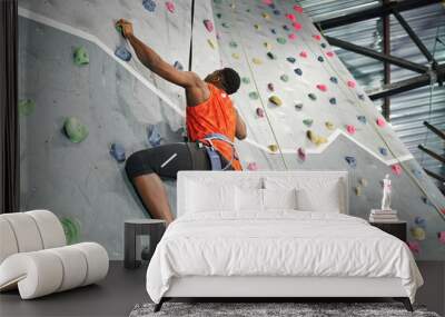 muscular african american man with alpine harness and safety rope climbing up bouldering wall Wall mural