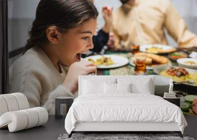 Muslim girl eating pilaf near blurred parents and ramadan dinner. Wall mural